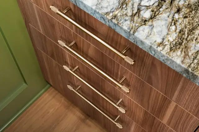 Close-up of walnut drawer fronts with long brass pulls beside an olive green cabinet and a granite counter